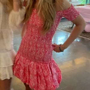 Rosa offshoulder kortklänning - Superfin rosa kortklänning med vitt mönster och offshoulder-modell. Klänningen har volang nertill och smockad överdel som ger en snygg passform. Perfekt för sommarens alla tillfällen och ger en fräsch och trendig look.