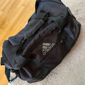 Svart träningsväska från Adidas - Stor svart träningsväska från Adidas med klassisk logga på sidan. Väskan har flera fack med dragkedja, bred axelrem och handtag med vaddering. Tillverkad i slitstark polyester och perfekt för gymmet eller sportaktiviteter.