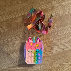 Regnbågsfärgad enhörningsväska pop it - Supergullig axelväska i form av en enhörning med regnbågsfärger och pop it-bubblor på framsidan. Väskan har rosa dragkedja och en färgglad axelrem. Perfekt för att bära småsaker och ha kul med pop it-effekten.