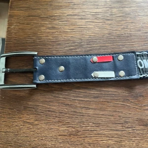 Tommy Hilfiger svart skärp med nitar - Svart bälte från Tommy Hilfiger med tydlig logga längs hela bandet. Skärpet har silverfärgat spänne, dekorativa nitar och detaljer i rött och silver. Materialet är skinn och textil, med vita kontrastsömmar för en cool look.