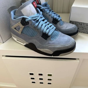 Nike Air Jordan 4 University Blue 42.5 - Nike Air Jordan 4 University Blue med ljusblå mocka, svarta och grå detaljer samt specklad häl. Klassisk siluett med snörning, meshpaneler och Jumpman-logga på baksidan. Perfekta för dig som vill sticka ut med en ikonisk sneaker.nypris 6499kr jag säljer dem för 2700kr