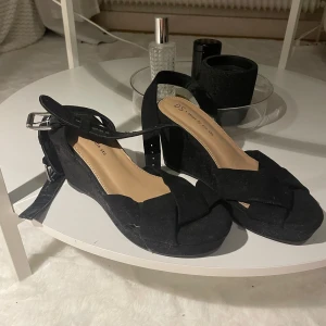 Svarta sandaler med kilklack från Din Sko - Snygga svarta sandaler från Din Sko med breda korslagda remmar i mocka och justerbart spänne vid ankeln. Skorna har en stabil kilklack och öppen tå, perfekt för att lyfta din sommarstil.  Storlek 36. Använda 1 gång 