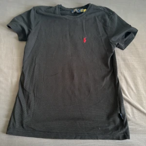Svart t-shirt från Polo Ralph Lauren - Svart t-shirt från Polo Ralph Lauren med klassisk röd logga broderad på bröstet. T-shirten är kortärmad och har en strukturerad bomullskänsla. Perfekt för dig som gillar stilrena och tidlösa plagg med en sportig touch.