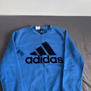Blå Adidas sweatshirt med logga - Säljer en blå sweatshirt från Adidas med stor svart logga framtill. Tröjan har rund hals, långa ärmar och är gjord i mjuk bomullsmix. Perfekt för en sportig och avslappnad stil. Klassisk passform och ribbade muddar vid ärmslut och nederkant.