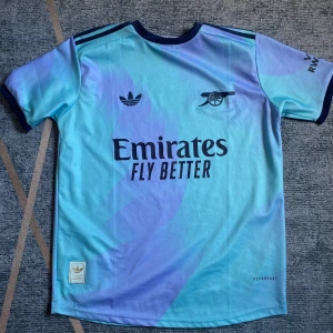 Arsenal x Adidas blå fotbollströja - Snygg Arsenal fotbollströja från Adidas i ljusblå och lila toner. Tröjan har korta ärmar, rund hals och tryck med 'Emirates Fly Better' samt klubbens kanon-logga. Materialet är lätt och andas, perfekt för match eller träning. Klassiska Adidas-ränder på axlarna.