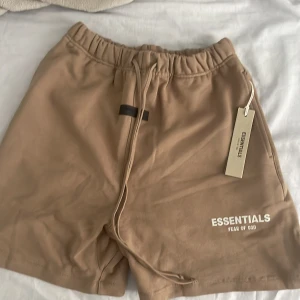 Beige shorts Essentials Fear of God - Snygga beige shorts från Essentials Fear of God med elastisk midja och långa snören framtill. Mjuk bomullskänsla och relaxed fit, perfekt för chill dagar. Diskret logga tryckt på benet och klassisk design utan mönster eller extra detaljer. Helt nya shorts utan användning.