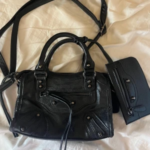 Svart handväska och plånbok - Säljer en ikonisk svart handväska i skinn med matchande plånbok. Väskan har både handtag och lång axelrem, coola metalldetaljer och dragkedjor framtill. Perfekt storlek för det viktigaste och ger en edgy vibe till din outfit.