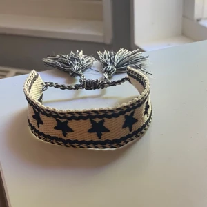 Beige och svart armband med stjärnor - Snyggt vävt armband i beige och svart med stjärnmönster runt om. Armbandet har justerbar knytning med två tofsar i ändarna för en cool boho-look. Perfekt accessoar för att lyfta din outfit med lite extra edge.