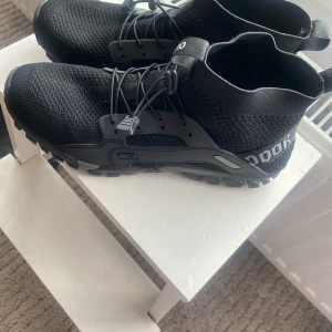 Svarta sneakers med grov sula - Svarta sneakers med stickad ovandel och grov, mönstrad sula för extra grepp. Skorna har snörning med elastiska snören och detaljer i syntet längs sidorna. Högre skaft med textdetalj och gul innersula. Perfekta för en sportig och urban stil. Helt nya!