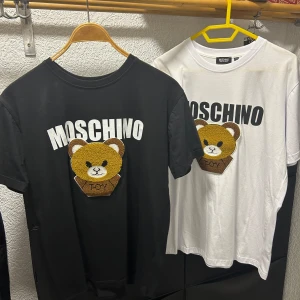 Moschino t-shirt med björn L - Säljer en Moschino t-shirt i storlek L, finns i både svart och vit. T-shirten har ett stort broderat björnmotiv och Moschino-logga framtill. Klassisk passform, rund hals och tillverkad i 100% bomull. Perfekt statement-plagg för dig som gillar streetstyle.