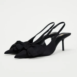 Svarta pumps med rosett från Zara - Stilrena svarta pumps från Zara med spetsig tå, smal klack och stor dekorativ rosett framtill. Skorna har öppen häl med smal rem och är tillverkade i ett matt tygmaterial som ger en elegant look. Perfekta för dig som vill sticka ut med en klassisk men trendig detalj. Knappt använda! 