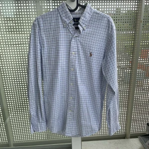 Rutig skjorta från Ralph Lauren - Säljer en snygg slim fit skjorta från Ralph Lauren i vitt och blått rutigt mönster. Skjortan har klassisk krage, knappar framtill och broderad logga på bröstet. Tillverkad i stretchig bomull för extra komfort. Perfekt för dig som gillar stilrena och tidlösa plagg.