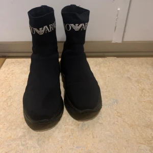  Emporio Armani - Svarta sockboots från Emporio Armani med högt skaft och tydlig logga i vitt upptill. Skorna har en chunky platt sula och är tillverkade i stretchigt syntetmaterial som sitter tight runt foten. Perfekta för en trendig streetstyle-look.