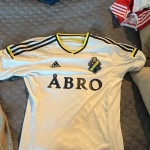 AIK vit matchtröja Adidas herr - AIK:s officiella vita matchtröja från Adidas med svart och gul detalj på krage och ärmslut. Klassiska svarta Adidas-ränder på axlarna, AIK:s klubbmärke broderat på bröstet och stor ÅBRO-logga framtill. Tillverkad i lätt och ventilerande polyester.