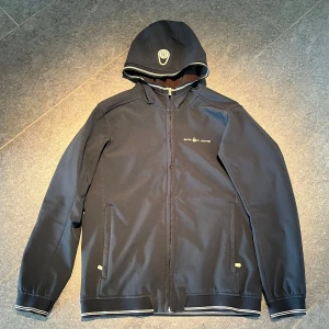 Sail racing jr grinder softshell hood  - En snygg vårjacka från sail racing i storlek 170, men passar S. Skicket är mycket bra, lite hår på insidan och lite slitage på texten framtill. Bredd: 50 cm, längd: 65 cm och armlängd: 59 cm. +/- 1 cm. Priset kan diskuteras! 