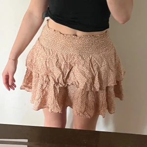 Beige/rosa volangkjol med mönster - Supersöt kort volangkjol i en persikofärg med vitt mönster. Kjolen har smock i midjan för en bekväm passform och flera lager volanger som ger en flowy känsla. 💘