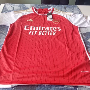 Arsenal hemmatröja Adidas röd/vit L - Arsenal hemmatröja från Adidas i storlek L. Röd med vita ärmar, guldiga detaljer och blixtmönster över hela tröjan. Broderat klubbmärke och Adidas-logga, samt sponsortryck på bröstet. Tillverkad i lätt och ventilerande Primegreen-material.