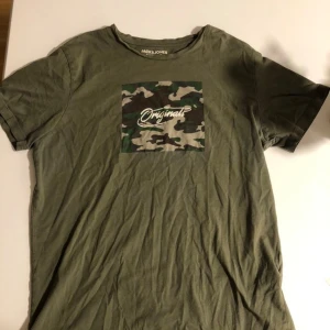 Grön camo t-shirt Jack & Jones - Snygg grön t-shirt från Jack & Jones Originals med camouflagemönstrad ruta och vit text på bröstet. Klassisk rund hals och korta ärmar. Skön bomullskvalitet som passar perfekt till jeans eller shorts.