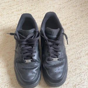 Svarta Nike Air Force 1 sneakers - Klassiska helsvarta Nike Air Force 1 sneakers i skinn med lågt skaft och rund tå. Skorna har snörning och diskret Nike-logga på sidan och bak. Perfekta för dig som gillar en clean och tidlös streetstil.