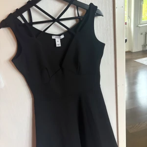 Svart kort klänning med korsade band - Snygg svart kort klänning från Oneness med korsade band i ryggen och figurnära överdel. Klänningen har ärmlös design och en lätt utställd kjol som ger en siluett. 