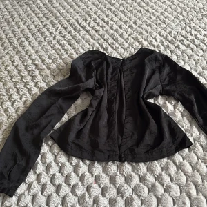 Svart blus från Cubus med öppen rygg - Svart blus från Cubus i storlek 36. Blusen har lång ärm, rund halsringning och knäppning bak med två små knappar, ryggen är också öppen. Materialet har en lätt glansig sidenkänsla och ger en elegant vibe. Perfekt att styla med jeans eller kjol för en snygg look.