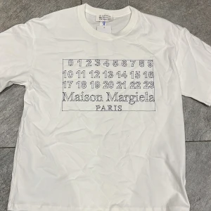 Maison Margiela vit t-shirt  - Vit t-shirt från Maison Margiela med ikoniskt svart siffertryck och logga på bröstet. Klassisk rund halsringning och korta ärmar. Tillverkad i mjuk bomull som känns skön mot huden. Perfekt för dig som gillar minimalistisk och stilren design. Vid fundering är det bara att skicka dm :)