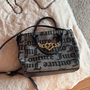 Juicy Couture handväska med kedjedetalj - Snygg handväska från Juicy Couture i svart och grått med stort logomönster. Väskan har svarta handtag, axelrem och en guldig kedja med hjärtdetalj framtill. Tillverkad i textil och skinnimitation, perfekt för dig som gillar statement-accessoarer.