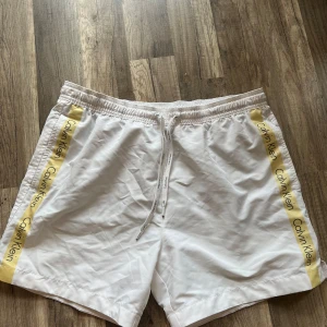 Vita badshorts Calvin Klein M - Fräscha vita badshorts från Calvin Klein med gula sidor och logga längs benen. Shortsen har elastisk midja med snörning och innerbyxa i mesh. Perfekta för stranden eller poolen, riktigt clean look och snygga detaljer.