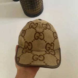 Gucci keps - Snygg keps från Gucci i beige och brunt med det klassiska GG-monogrammet över hela kepsen. Justerbar rem baktill i brunt läder och detaljer i guld. Tillverkad i canvas med läderdetaljer. Perfekt för dig som vill sticka ut med en lyxig accessoar. Ganska använd ser fortforande bra ut 
