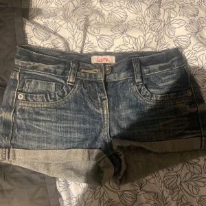 Blå jeansshorts från Sophie - Snygga blå jeansshorts från Sophie med slitna detaljer och uppvikta benslut. Klassisk femficksmodell med bälteshällor och knappstängning. Perfekta för varma dagar och en avslappnad look. midjemått är 31