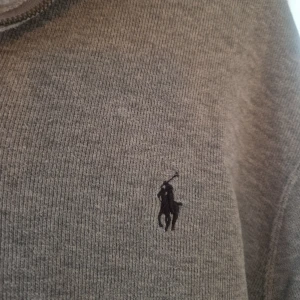 Grå half zip tröja Polo Ralph Lauren - Snygg grå långärmad tröja från Polo Ralph Lauren med half zip och klassisk broderad logga på bröstet. Tröjan är i mjuk bomull och har en avslappnad passform, perfekt till jeans eller chinos. Enkel och stilren design som funkar året runt.