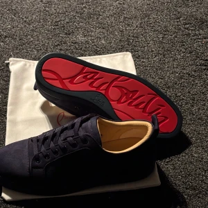 Mörkblå sneakers från Christian Louboutin - Säljer ett par mörkblå sneakers från Christian Louboutin med klassisk röd sula och diskret logga på sidan. Skorna har snörning och är tillverkade i mocka med brodering på baksidan. Perfekta för dig som vill ha något stilrent men ändå sticker ut. Priset kan diskuteras