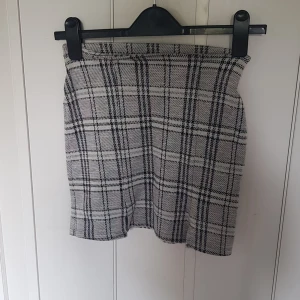 Rutig kortkjol från Lindex - Superfin rutig kortkjol från Lindex i grått, svart och vitt. Kjolen har en liten slits framtill och är i mjukt, stretchigt material. Perfekt för dig som gillar klassisk stil med en twist.