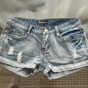 Ljusblå jeansshorts med slitningar - Snygga low waist ljusblå jeansshorts i storlek xs/s från Fashion& Jeans 2013 med slitna detaljer både fram och bak. Klassisk femficksmodell, uppvikta benslut och normal passform. Perfekta för varma dagar när du vill ha en avslappnad och trendig look.