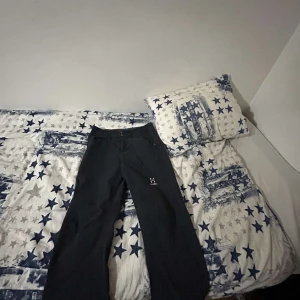 Svarta täckbyxor från Haglöfs - Svarta täckbyxor från Haglöfs med dragkedjor vid fickorna och logga på låret. Byxorna har normal passform och är tillverkade i slitstarkt syntetmaterial, perfekta för vinterns alla äventyr.