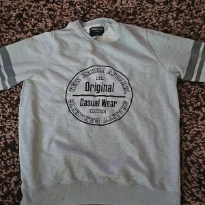 Grå sweatshirt från Identic med tryck - Grå sweatshirt från Identic med svart tryck på bröstet där det står 'Original Casual Wear'. Tröjan har långa ärmar med två mörkgrå ränder på varje ärm och rund halsringning. Perfekt för en chill och avslappnad stil.