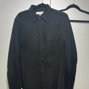 Svart manchesterskjorta Studio Total - Svart långärmad skjorta i manchester från Studio Total. Klassisk krage med knappar och en bröstficka. Skjortan har en stilren och enkel look som passar perfekt till jeans eller chinos.