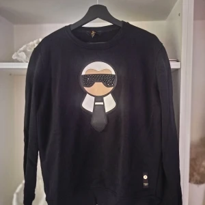 Svart Fendi sweatshirt med Karl Lagerfeld-motiv - Svart sweatshirt från Fendi med ikoniskt Karl Lagerfeld-motiv framtill, dekorerat med svarta pärlor på glasögonen. Tröjan har rund halsringning och långa ärmar. Perfekt för dig som gillar statement-plagg och exklusiva designer.