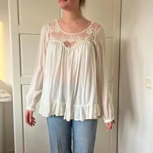 Vit blus med spets och volang - Romantisk vit blus med transparent spets upptill och fina broderade detaljer. Långärmad modell med volangkant nertill och knappar i ryggen. Luftig och flowy passform, perfekt för dig som gillar en söt och bohemisk stil.