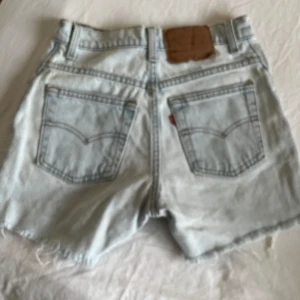Levi's ljusblå jeansshorts med fransar - Ljusblå vintag jeansshorts från Levi's med klassiska fem fickor, råa fransiga benslut och hög midja. Snyggt slitna detaljer och ikonisk läderpatch bak. Perfekta för en chill sommarlook.