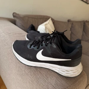 Svarta Nike sneakers med vit sula - Svarta Nike sneakers med vit sula och klassisk vit swoosh på sidan. Ovandelen är i mesh för bra andningsförmåga och skon har snörning. Perfekta för träning eller vardagsstil, med en sportig och clean look.