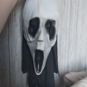 Scream maskeradmask med svart tyg  - Säljer en ikonisk Scream-mask med vit och svart ansiktsform i plast och svart tyg som täcker bakhuvudet. Perfekt för maskerad eller halloween. Masken har de klassiska utsträckta ögon- och munöppningarna och är lätt att bära tack vare det mjuka tyget baktill.