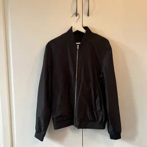 Svart bomberjacka från J.Lindeberg - Svart bomberjacka från J.Lindeberg i klassisk modell med dragkedja framtill! Använd fåtal gånger                               Storlek: M 