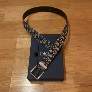 Svart och beige Dior bälte med monogram - Säljer ett snyggt Dior bälte med klassiskt monogrammönster i svart och beige. Bältet har en silverfärgad metallspänne med Dior-logga och insida i svart skinn. Perfekt accessoar för att lyfta din outfit med lyxig känsla.