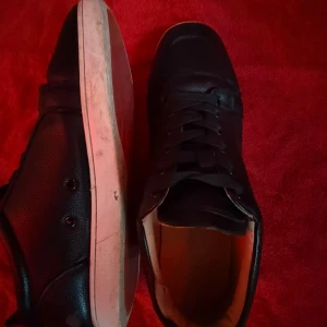 Svarta sneakers från Louboutin - Snygga svarta sneakers från Louboutin med klassisk röd sula och diskret logga undertill. Skorna har snörning, rund tå och är tillverkade i skinn med vit kant runt sulan. Perfekta för dig som vill ha en stilren och exklusiv look.