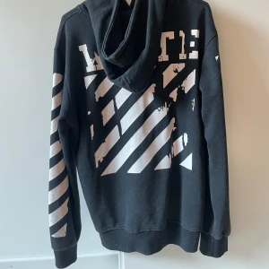 Off-White svart hoodie med tryck - Svart hoodie från Off-White med ikoniska vita diagonala ränder på ärmar och rygg. Framsidan har ett  tryck i vitt och gult samt texten 'WHITE' nertill. Oversized passform och huva, tillverkad i mjuk bomull. 
