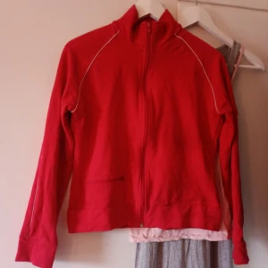 Röd zip-jacka med vita detaljer - Röd jacka med hel dragkedja och vita detaljer längs axlar och ärmar. Hög krage och långa ärmar, perfekt för en sportig look. Jackan har fickor framtill och är tillverkad i mjukt bomullsmaterial.