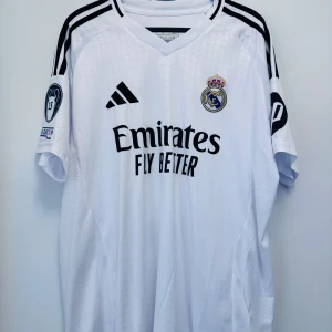Real Madrid Bellingham Fan edition storlek L - Real Madrid fotbollströja Tryck med Bellingham och nummer 5 på ryggen.  Champions League-märke på ärmen. Trycket under blev felplacerat så här köpt en ny säljer drf denna billigt. Jag skickar ingenting så får mötas upp. Liljeholmen/gröndal, Slussen/TC. Har mer tröjor så fråga gärna om  det är någon annan du skulle vilja köpa.   Tar Swish och kontanter. 