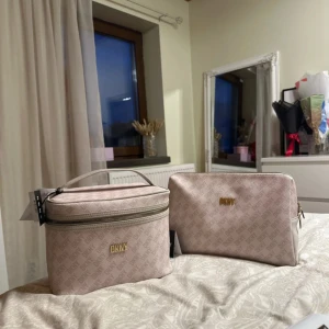 Beige necessär- och sminkväska från DKNY - Två snygga väskor från DKNY i beige med diskret logomönster. Den ena är en boxig necessär med handtag och dragkedja, den andra en sminkväska med guldig logga och dragkedja. Båda i tåligt syntetmaterial, perfekta för att organisera dina skönhetsprylar. Båda kostar 900kr tillsammans