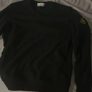 Svart Stone Island sweatshirt - Svart sweatshirt från Stone Island med klassisk rund hals och ikonisk logotyp-patch på vänster ärm. Tröjan har en clean look och är tillverkad i mjuk bomull, perfekt för en stilren och avslappnad vibe.
